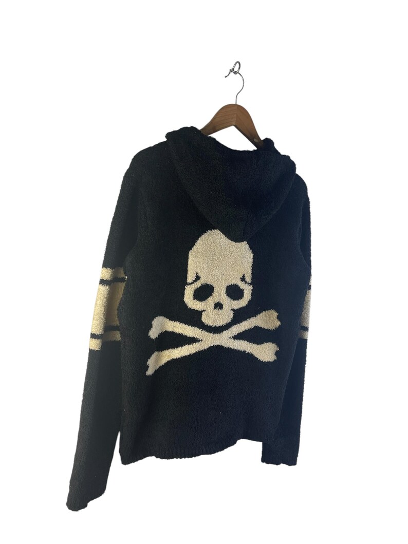 May include: Black hooded sweater with a skull and crossbones design on the back. The sweater has gold stripes on the sleeves and a matching gold skull graphic. The sweater is hanging on a wooden hanger.