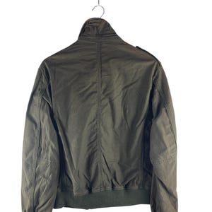 May include: Olive green bomber jacket with a ribbed collar, cuffs, and hem. The jacket has a full zipper closure and a stand-up collar. The back of the jacket is visible, showing the seams and construction details. The jacket is hanging on a metal hanger.