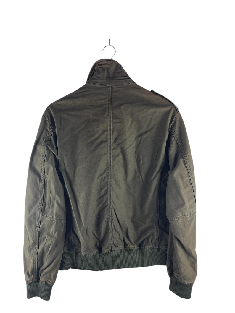 May include: Olive green bomber jacket with a ribbed collar, cuffs, and hem. The jacket has a full zipper closure and a stand-up collar. The back of the jacket is visible, showing the seams and construction details. The jacket is hanging on a metal hanger.