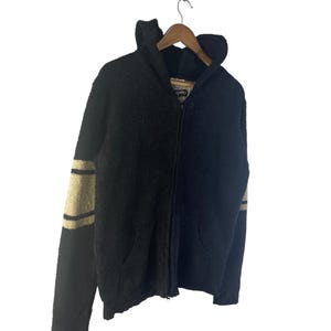May include: Black zip-up hooded sweater with tan stripes on the sleeves. The sweater has a textured, fuzzy appearance, a hood, and two front pockets. It is hanging on a wooden hanger against a white background.