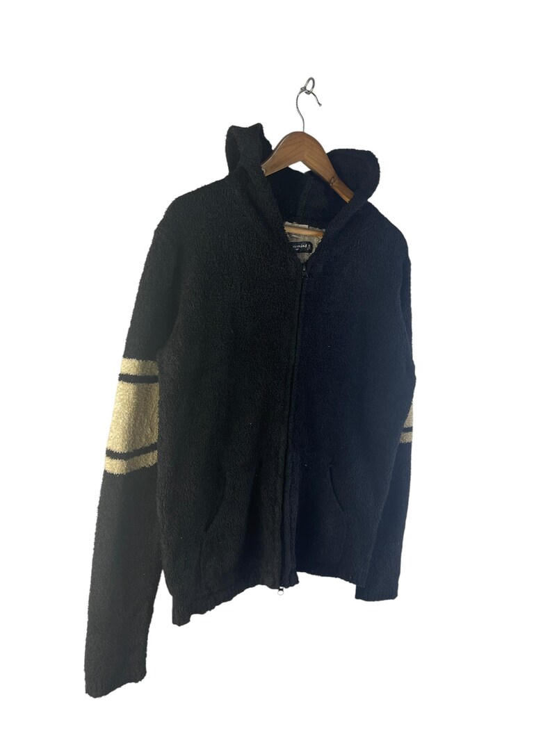 May include: Black zip-up hooded sweater with tan stripes on the sleeves. The sweater has a textured, fuzzy appearance, a hood, and two front pockets. It is hanging on a wooden hanger against a white background.