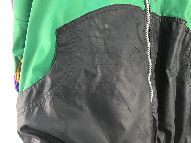 Puede incluir: Una chaqueta negra, verde y morada con cierre de cremallera. La chaqueta tiene un bolsillo en el lado izquierdo.