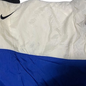 Op de afbeelding: Een wit en blauw nylon jack met een zwart Nike swoosh-logo aan de bovenkant links. Het jack heeft een ritssluiting en een blauwe voering.