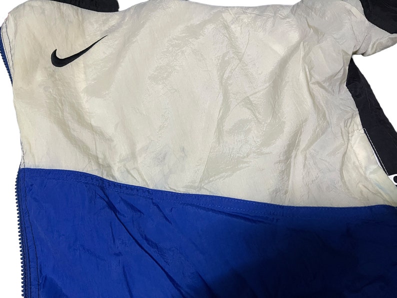 Op de afbeelding: Een wit en blauw nylon jack met een zwart Nike swoosh-logo aan de bovenkant links. Het jack heeft een ritssluiting en een blauwe voering.