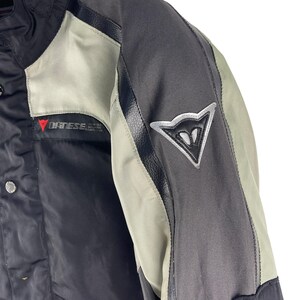 K&ouml;nnte beinhalten: Nahaufnahme einer schwarzen und grauen Motorradjacke mit einem silbernen Logo auf dem &Auml;rmel. Die Jacke hat hellgraue Eins&auml;tze und der Markenname "DAINES" ist sichtbar. Die Jacke scheint aus strapazierf&auml;higen, wetterfesten Materialien zu bestehen.