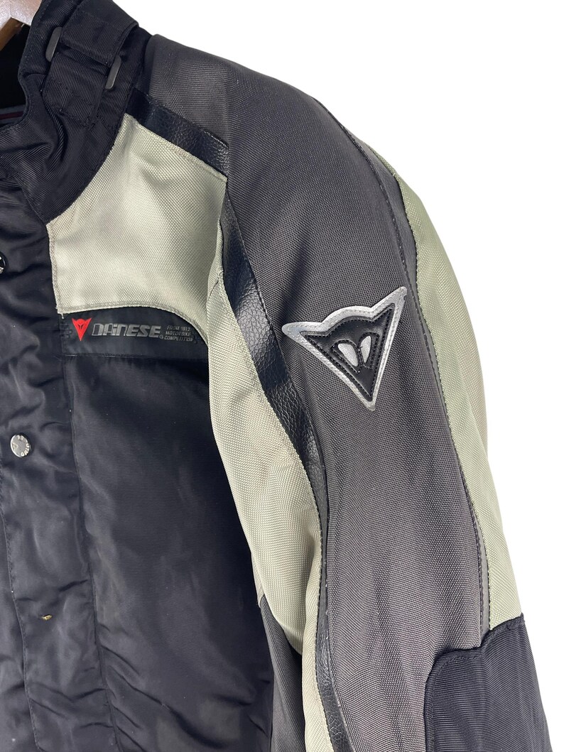 K&ouml;nnte beinhalten: Nahaufnahme einer schwarzen und grauen Motorradjacke mit einem silbernen Logo auf dem &Auml;rmel. Die Jacke hat hellgraue Eins&auml;tze und der Markenname "DAINES" ist sichtbar. Die Jacke scheint aus strapazierf&auml;higen, wetterfesten Materialien zu bestehen.