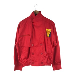 May include: A red jacket with a double-breasted closure and a patch on the right side that says "KYGNUS Ba.n RACING SPORTS 98-99".