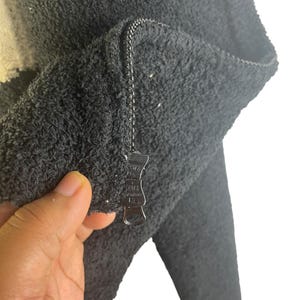May include: Close-up of a dark gray, textured jacket with a zipper. The jacket appears to be made of a soft, plush material. The zipper is silver and has a unique pull.