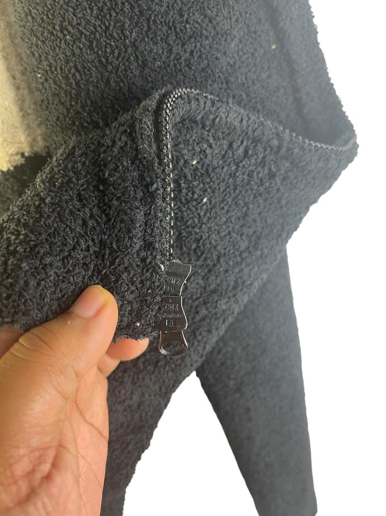 May include: Close-up of a dark gray, textured jacket with a zipper. The jacket appears to be made of a soft, plush material. The zipper is silver and has a unique pull.