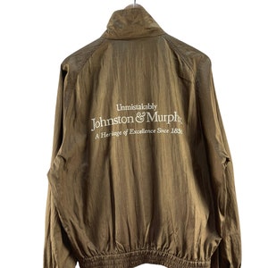 Puede incluir: Una chaqueta marrón con cierre de cremallera y un logotipo bordado en blanco que dice "Unmistakably Johnston & Murphy. A Heritage of Excellence Since 1850."