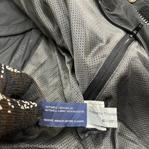 Puede incluir: Una chaqueta negra de nylon con forro de malla. La etiqueta dice "OUTSHELL: 100% NYLON OUTSHELL LINING: 100% POLYESTER REMOVE ARMOUR BEFORE CLEAKING" y "Produced For Triumph All rights reserved".