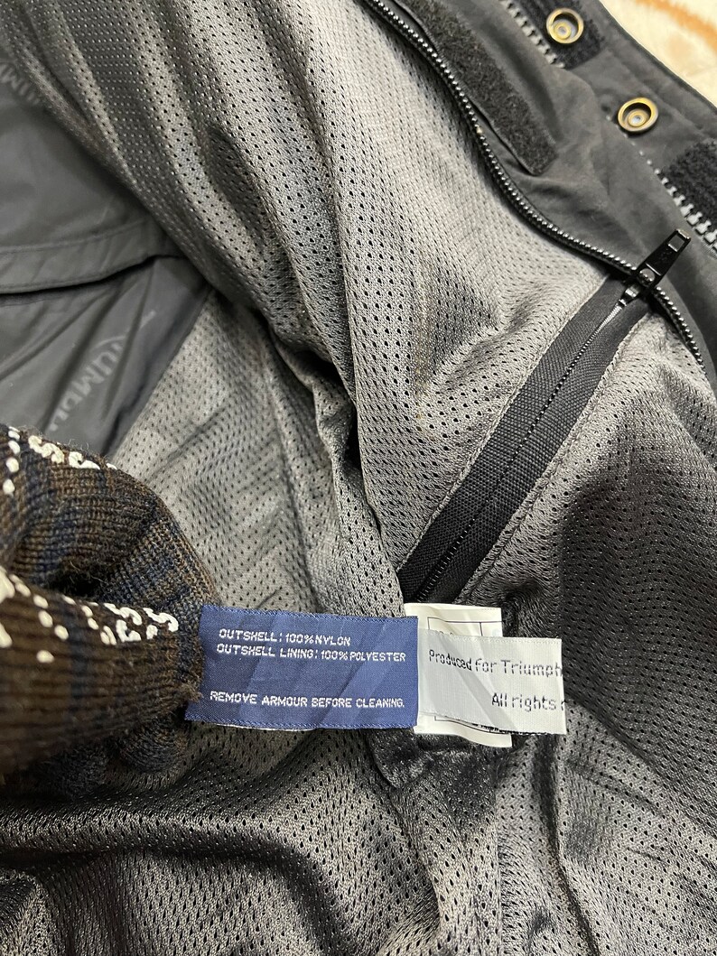 Puede incluir: Una chaqueta negra de nylon con forro de malla. La etiqueta dice "OUTSHELL: 100% NYLON OUTSHELL LINING: 100% POLYESTER REMOVE ARMOUR BEFORE CLEAKING" y "Produced For Triumph All rights reserved".