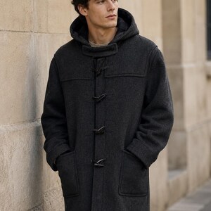 May include: A charcoal gray wool duffle coat featuring a hood, long sleeves, and a front closure with three black toggle buttons. The coat includes two large patch pockets on the front and reaches mid-thigh length with a structured, straight fit.