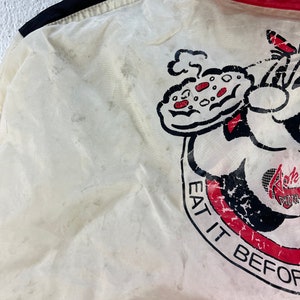Puede incluir: Chaqueta blanca con ribete rojo y negro. La chaqueta tiene un personaje de dibujos animados sosteniendo una pizza con el texto "Eat it before it gets cold" y "Ralph's Pizza" en ella.