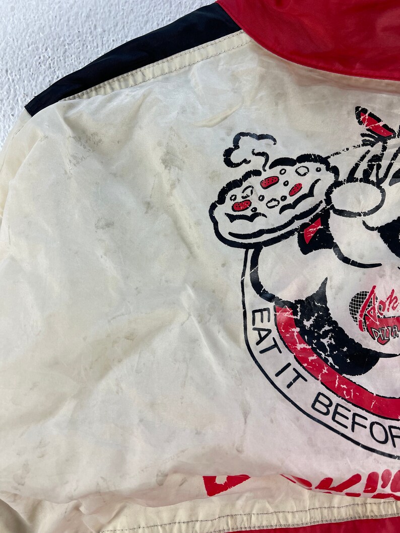 Puede incluir: Chaqueta blanca con ribete rojo y negro. La chaqueta tiene un personaje de dibujos animados sosteniendo una pizza con el texto "Eat it before it gets cold" y "Ralph's Pizza" en ella.
