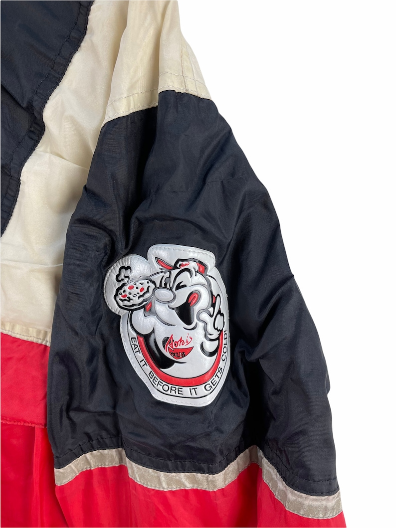 Puede incluir: Chaqueta negra y roja con una raya blanca y negra. La chaqueta tiene un parche grande en la manga izquierda con un personaje de dibujos animados sosteniendo una pizza y el texto "Roteli's Pizza" y "Eat it before it gets cold".