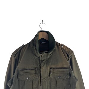 May include: Olive green military-style jacket with a high collar and multiple pockets. The jacket features a zipper closure, epaulets on the shoulders, and a ribbed collar. The jacket is hanging on a wooden hanger against a white background.