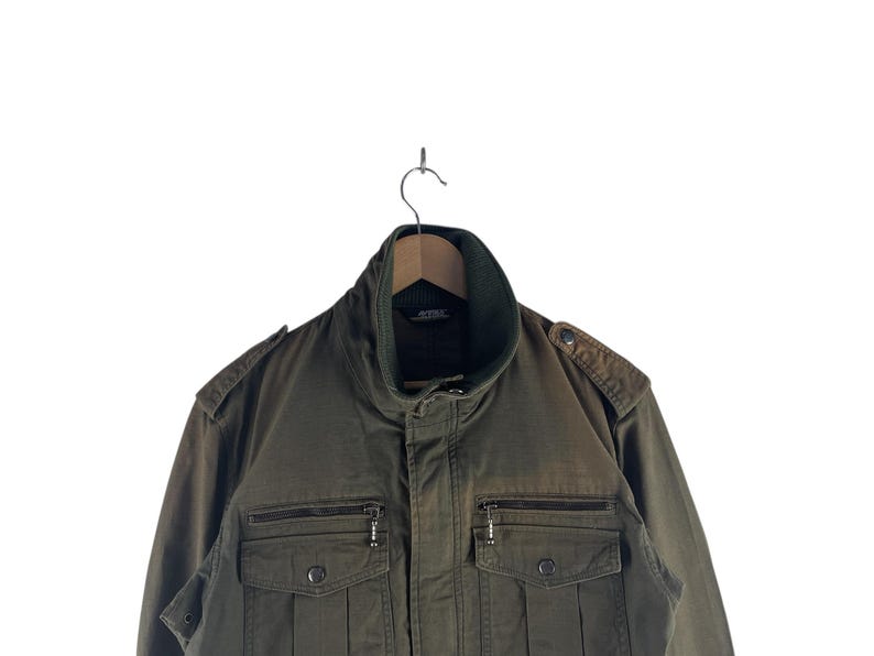 May include: Olive green military-style jacket with a high collar and multiple pockets. The jacket features a zipper closure, epaulets on the shoulders, and a ribbed collar. The jacket is hanging on a wooden hanger against a white background.