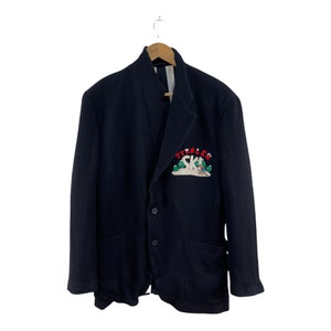 May include: A black blazer with a ribbed texture and a patch on the left breast that says "Tyrolean Ski" in red and green lettering. The patch features a cartoon skier in a green hat and red pants.