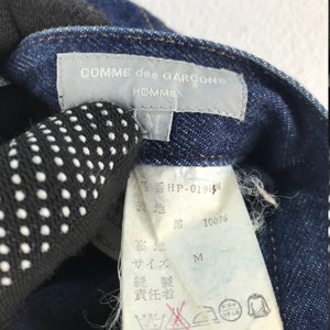 以下が含まれることがあります： 「コムデギャルソン オム」と「品番 HP-0190M」の白い文字が書かれた青いデニムのタグのクローズアップ。タグには日本語でのお手入れ方法も記載されています。