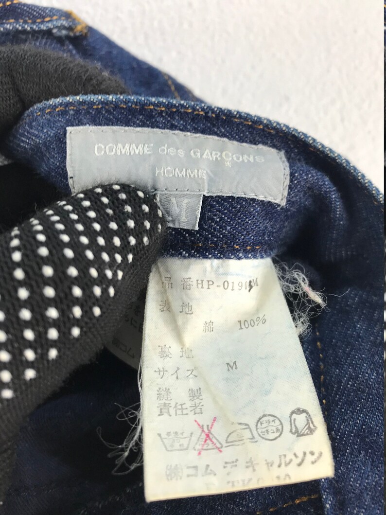 以下が含まれることがあります： 「コムデギャルソン オム」と「品番 HP-0190M」の白い文字が書かれた青いデニムのタグのクローズアップ。タグには日本語でのお手入れ方法も記載されています。