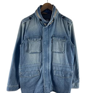 Puede incluir: Chaqueta vaquera azul claro con cuello con cremallera y m&uacute;ltiples bolsillos delanteros. La chaqueta tiene cierre de botones y charreteras en los hombros. Cuelga de una percha de madera sobre un fondo blanco.