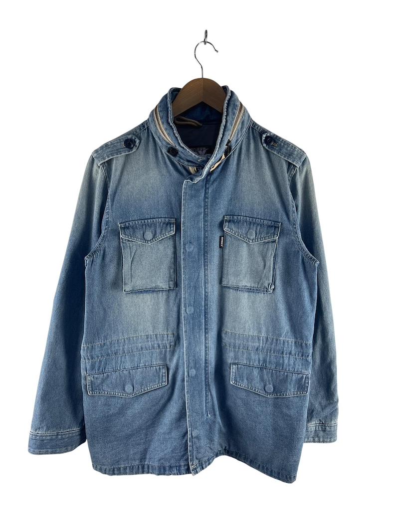 Puede incluir: Chaqueta vaquera azul claro con cuello con cremallera y m&uacute;ltiples bolsillos delanteros. La chaqueta tiene cierre de botones y charreteras en los hombros. Cuelga de una percha de madera sobre un fondo blanco.