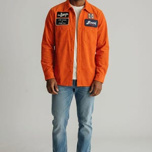 Puede incluir: Una camisa naranja abotonada con parches y vaqueros azul claro. La camisa tiene dos bolsillos en el pecho y una camiseta interior blanca. Los parches tienen texto y logotipos. La persona lleva zapatillas blancas.
