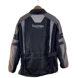 Puede incluir: Chaqueta de moto negra y gris con la palabra "TRIUMPH" en letras blancas en la espalda.