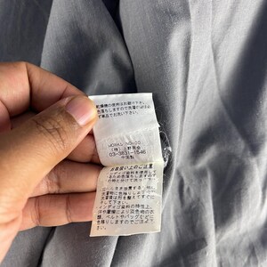 Puede incluir: Primer plano de una prenda gris con una etiqueta blanca. La etiqueta tiene texto japon&eacute;s y la marca "WORKS NO-00". La etiqueta tambi&eacute;n incluye instrucciones de cuidado. La prenda est&aacute; hecha de una tela suave y texturizada.