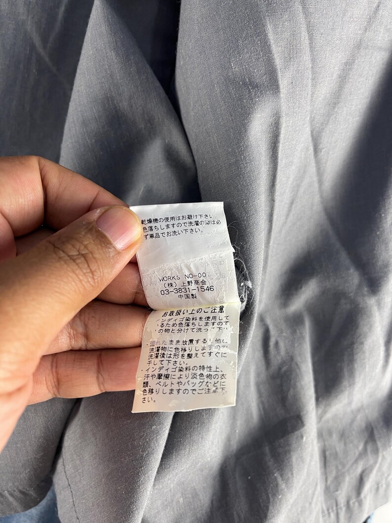 Puede incluir: Primer plano de una prenda gris con una etiqueta blanca. La etiqueta tiene texto japon&eacute;s y la marca "WORKS NO-00". La etiqueta tambi&eacute;n incluye instrucciones de cuidado. La prenda est&aacute; hecha de una tela suave y texturizada.