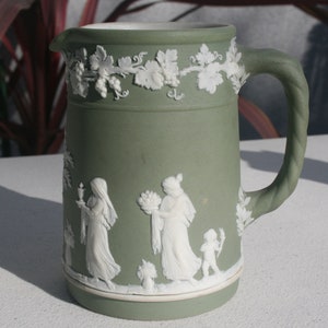 May include: A green ceramic pitcher with white raised relief designs. The pitcher has a handle and a spout. The design features a vine with leaves and grapes, and three figures in a scene.