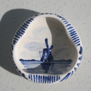 Puede incluir: Un plato de cerámica blanco con un diseño de molino de viento azul y blanco. El plato tiene un borde festoneado y un borde rayado azul y blanco.