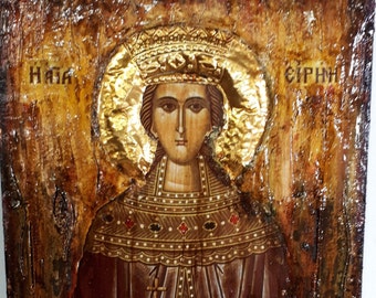 Puede incluir: Un icono de estilo vintage que representa a una mujer con una corona y una larga túnica oscura. El icono está pintado en un panel de madera con fondo dorado y tiene un aspecto desgastado y antiguo. La mujer sostiene una cruz en su mano derecha y su mano izquierda está levantada en un gesto de bendición. El icono está etiquetado como "Haya" y "Eirini" en escritura cirílica.