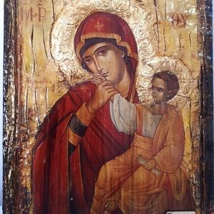 Può includere: Un'icona dipinta a mano che rappresenta la Vergine Maria che tiene in braccio il bambino Gesù. L'icona è dipinta su un pannello di legno con sfondo dorato e presenta un'aureola rossa e dorata intorno alla testa della Vergine Maria. L'icona è incorniciata in una cornice di legno scuro.