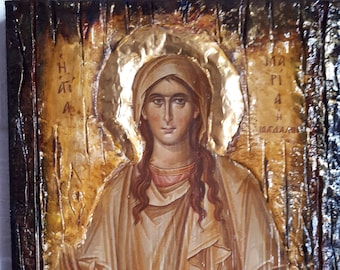 May include: A religious icon painting of a woman holding a pitcher. The woman is wearing a brown robe and has long brown hair. The background is gold and the icon is framed in a dark wood frame. The text on the icon reads "Η ΑΓΙΑ ΠΑΡΑΣΚΕΥΗ".