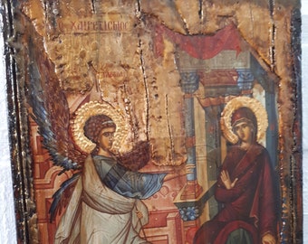 Op de afbeelding: Een religieus icoon schilderij in vintage stijl dat de Annunciatie voorstelt. De engel Gabriël, met vleugels, is afgebeeld in een witte robe met blauwe accenten, en steekt een hand uit naar de Maagd Maria, die op een troon zit. Het icoon is geschilderd op een houten paneel met een verweerde, oude uitstraling.