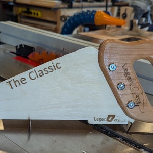 May include: A white wooden saw with a wooden handle. The saw is labelled "The Classic" and "Legendary" on the blade and handle. The saw is sitting on a table saw.