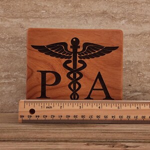 Op de afbeelding: Een houten plaquette met een zwart gegraveerd medisch symbool en de letters "PA". De plaquette is 10 cm breed en 10 cm hoog.