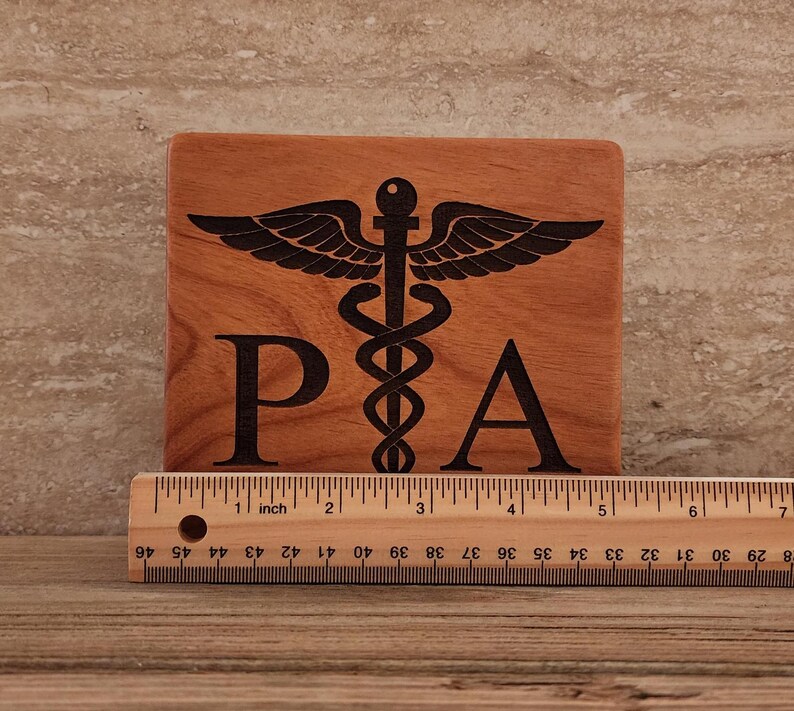Op de afbeelding: Een houten plaquette met een zwart gegraveerd medisch symbool en de letters "PA". De plaquette is 10 cm breed en 10 cm hoog.