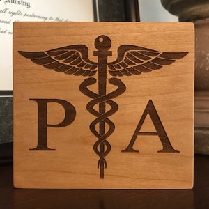 Op de afbeelding: Een houten plaquette met een bruine achtergrond en een lasergegraveerd medisch symbool, de staf van Asklepios, met de letters "PA" aan weerszijden van het symbool.