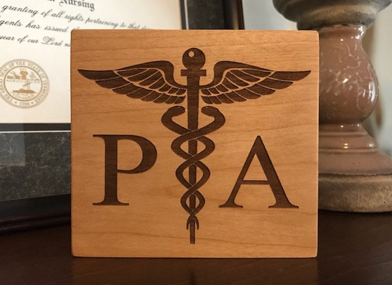 Op de afbeelding: Een houten plaquette met een bruine achtergrond en een lasergegraveerd medisch symbool, de staf van Asklepios, met de letters "PA" aan weerszijden van het symbool.
