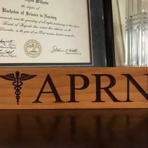 May include: A wooden sign with the text "APRN" and a medical symbol, a caduceus, with wings and a snake wrapped around a staff.