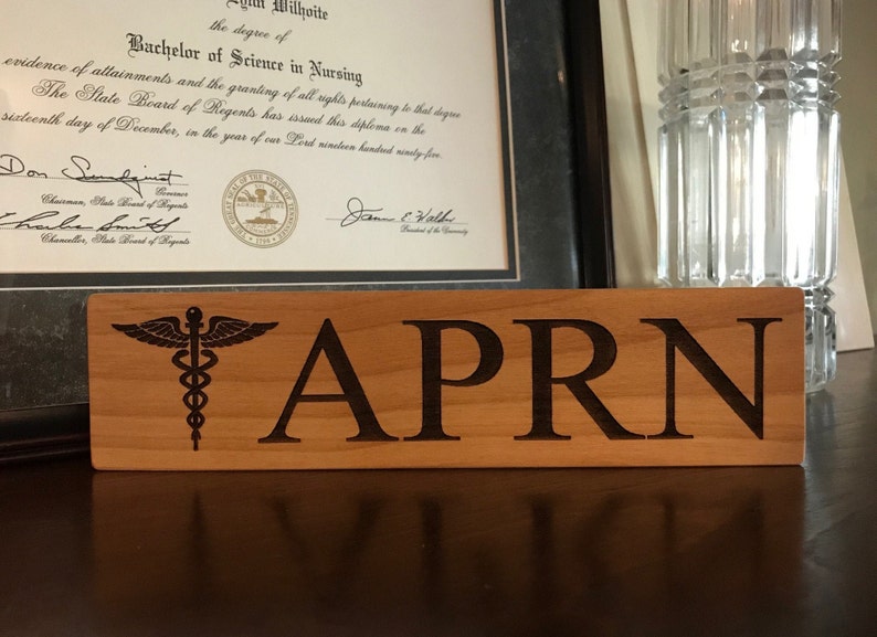 May include: A wooden sign with the text "APRN" and a medical symbol, a caduceus, with wings and a snake wrapped around a staff.