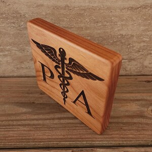 Op de afbeelding: Een houten plaquette met een donkerbruine vlek, met een zwart-wit gegraveerd medisch symbool met de letters "PA" eronder.