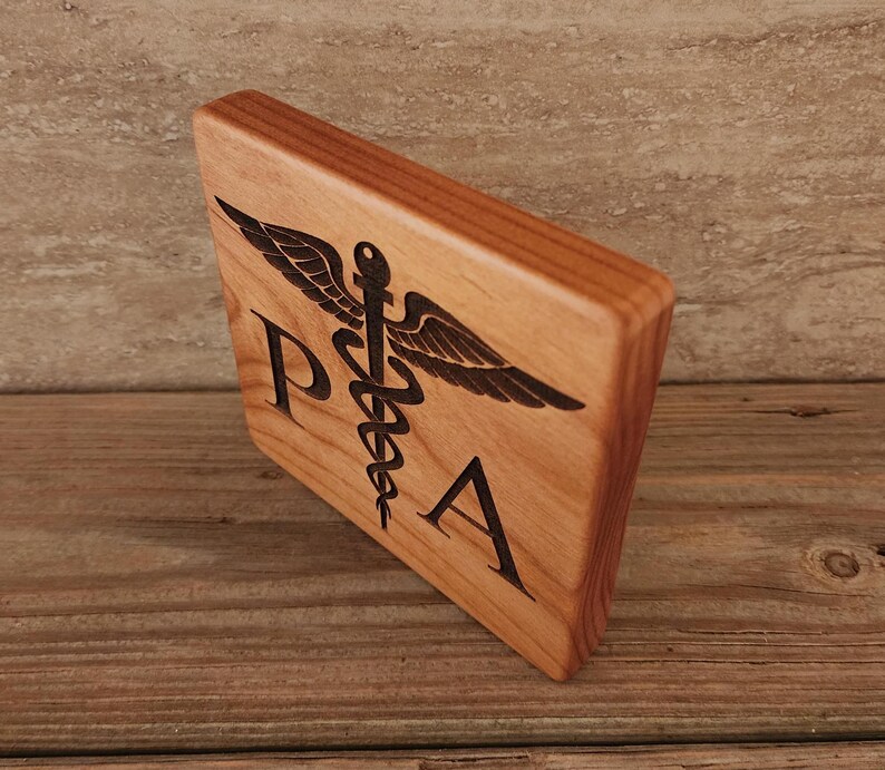 Op de afbeelding: Een houten plaquette met een donkerbruine vlek, met een zwart-wit gegraveerd medisch symbool met de letters "PA" eronder.