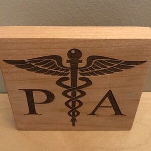 Op de afbeelding: Een houten blok met de letters "PA" en een gegraveerd medisch symbool.