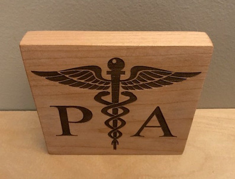 Op de afbeelding: Een houten blok met de letters "PA" en een gegraveerd medisch symbool.