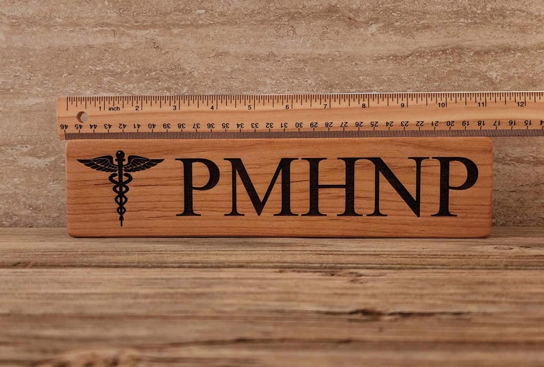 May include: A wooden sign with the text "PMHNP" and a medical caduceus symbol. The sign is made of light brown wood and has a natural finish.
