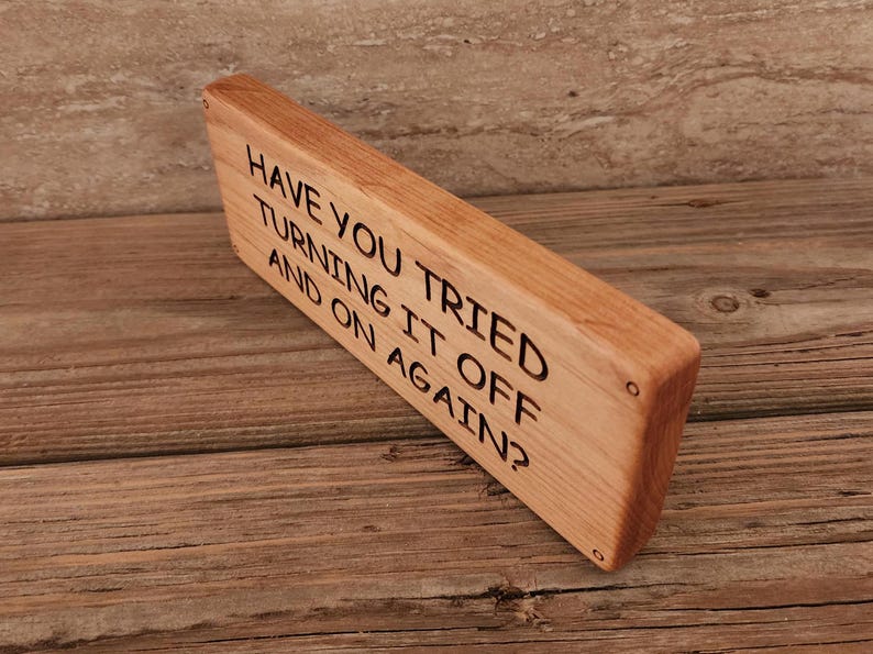 May include: A wooden sign with the text "Have you tried turning it off and on again?"  The sign is made of light brown wood and has a slightly distressed look.